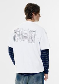 Witte katoenen t-shirt met een gedetailleerde grafiek op de achterkant, die de binnenkant van een vliegtuig weergeeft, met twee zittende figuren en een uitzicht uit het raam.