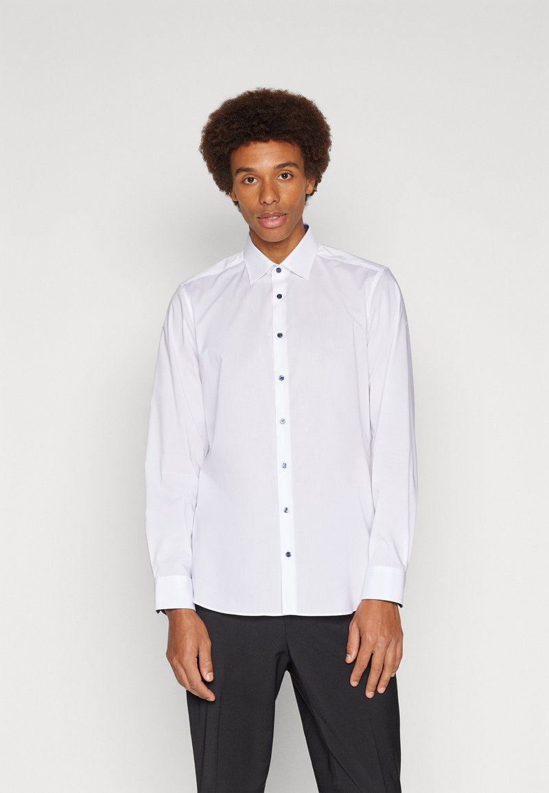 Chemise blanche à manches longues en tissu lisse avec un col classique, ornée de boutons noirs sur le devant et de poignets français. Coupe ajustée.