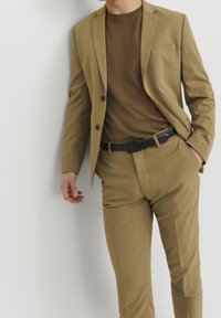 Beige kostym med en figursydd kavaj och byxor, tillsammans med en brun tröja. Har en ficka på kavajen, svart bälte, slät tyg.