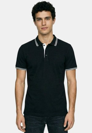 Jeune homme aux cheveux bouclés portant un polo noir à manches courtes avec une bordure blanche sur le col et les manches, debout devant un fond uni.