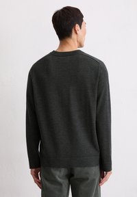 Mann mit kurzem dunkelhaar, der einen dunkelgrauen langärmligen Pullover und graue Cordhosen trägt, steht mit dem Rücken zur Wand vor einer weißen Wand.