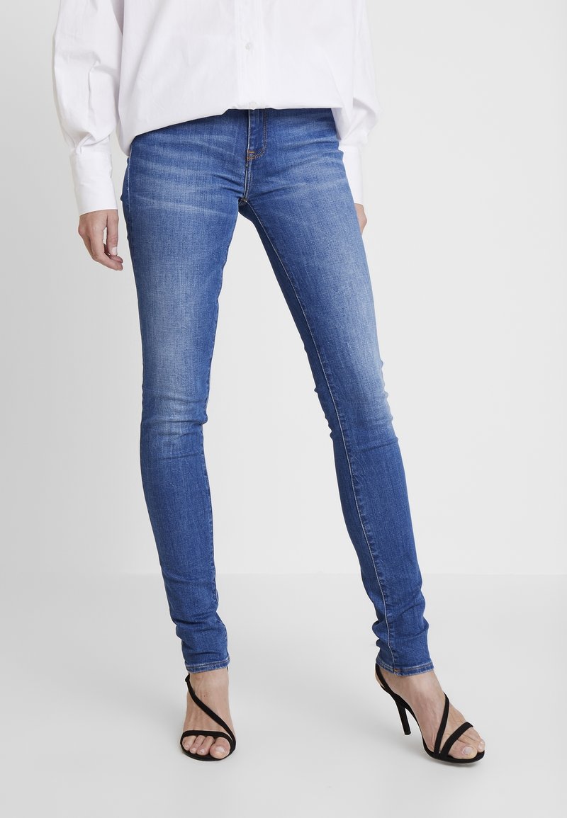 Jeans ajustados de mezclilla azul, con cintura de altura media, ligera decoloración y un ajuste ceñido hasta los tobillos, llevados con tacones negros de tiras.