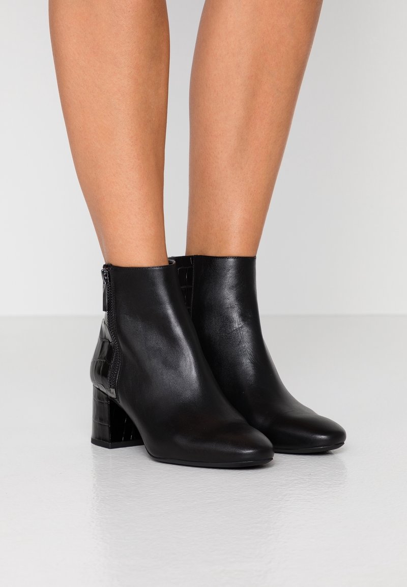 Jambes portant des bottines en cuir noir à talons blocs avec des fermetures éclair latérales, debout sur un sol blanc brillant.