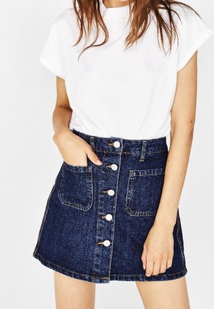 Vrouw draagt een wit shirt met korte mouwen, ingestopt in een donkerblauwe denim rok met knopen aan de voorkant en voorzakken.