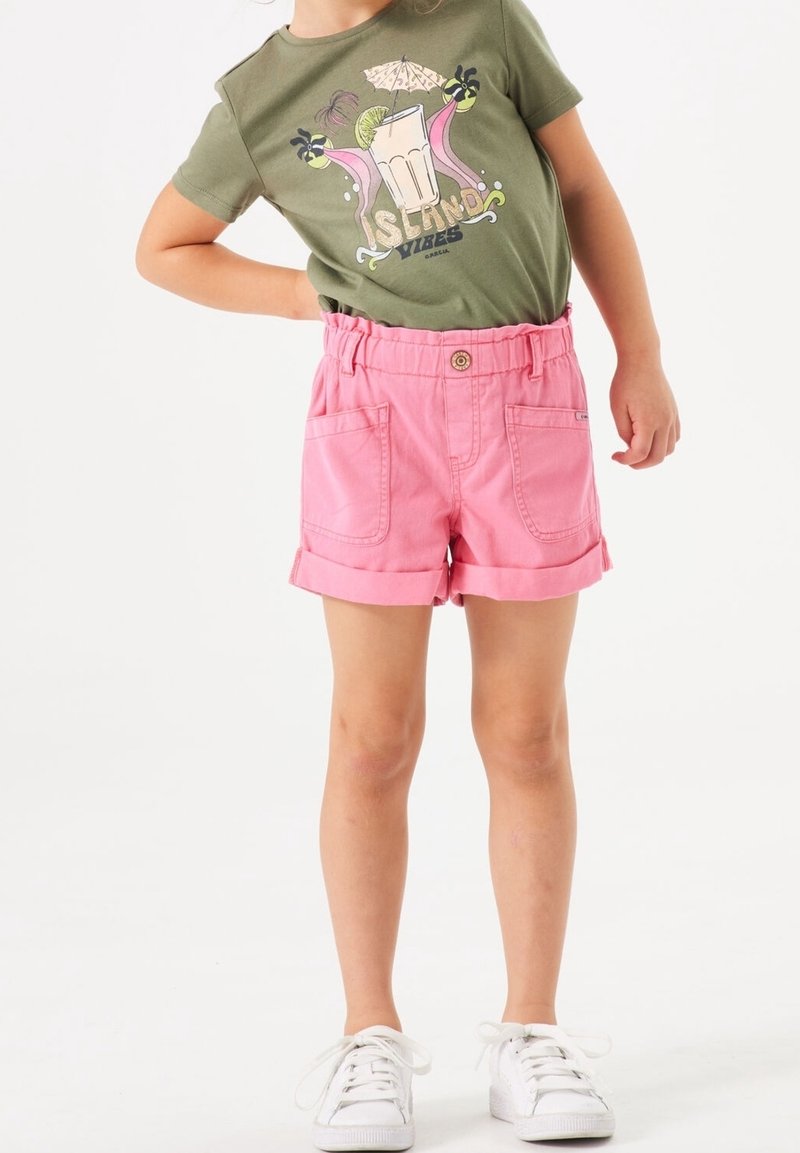 Shorts tissés roses avec un ourlet roulé, dotés de poches latérales et d'une taille élastique. Associés à un t-shirt graphique vert. Baskets blanches.