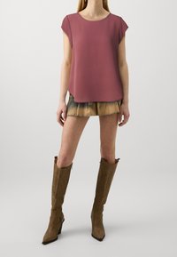 Top corto bordeaux a maniche corte, texture liscia, abbinato a una mini gonna a strati beige e denim e stivali marroni al ginocchio con punta affusolata.