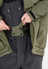 Outerwear mit einer olivgrünen Jacke und einer schwarzen Innenschicht. Bemerkenswerte Details sind Reißverschlüsse, ein Druckknopfverschluss und ein strukturiertes Gewebe.