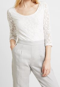 Vit spets långärmad topp med rundad halsringning, tillsammans med ljusgrå high-waisted linbyxor med veck framtill och sidofickor.
