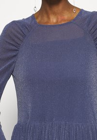 Lila mekko, jossa on pitkät hihat, ribbed rakenne ja hienovarainen kiilto; siinä on kerätty yksityiskohta olkapäillä ja liehuva hame.