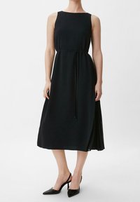 Nainen, joka pitää päällään hihattomaa mustaa midimekkoa, jossa on ohut vyö, ja mustia kärjellisiä slingback-korkokenkiä, seisoo valkoista taustaa vasten.