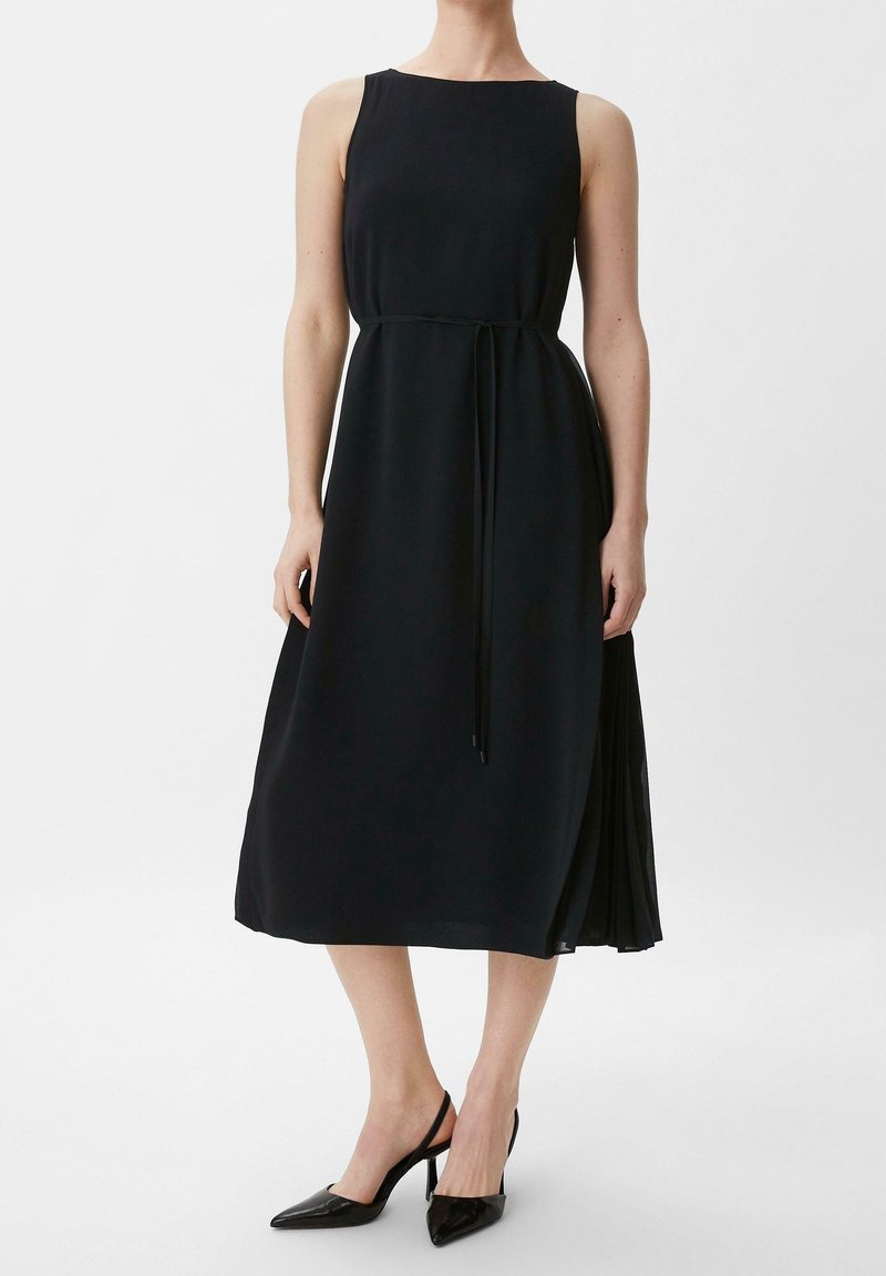 Nainen, joka pitää päällään hihattomaa mustaa midimekkoa, jossa on ohut vyö, ja mustia kärjellisiä slingback-korkokenkiä, seisoo valkoista taustaa vasten.