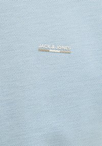 Tissu bleu clair texturé avec un motif tissé subtil. Présente une plaque de logo argentée portant l'inscription « JACK & JONES » près du haut.