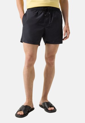 Mann trägt schwarze Badeshorts mit Kordelzug, schwarze Sandalen mit Kreuzriemen und ein gelbes Hemd, steht mit entspannten Händen seitlich am Körper.