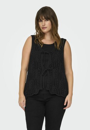 Schwarze gestrickte Weste mit offenem Vorderteil, die ein Spitzenmuster und eine Bindeschließe aufweist. Getragen über einem figurbetonten schwarzen Oberteil, kombiniert mit schwarzen Hosen.