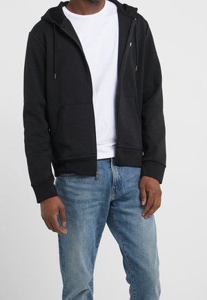 Man wearing a black zip-up hoodie over a white t-shirt and light blue jeans, standing against a plain background.