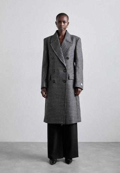 Manteau gris à carreaux pied-de-poule croisé avec larges revers et poches latérales, doté d'une silhouette structurée et d'une coupe ajustée. Associé à un pantalon noir large.