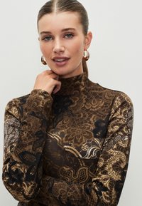 Brown turtleneck top with intricate black and gold lace patterns. The fabric appears smooth and fitted, featuring long sleeves and a high neckline.