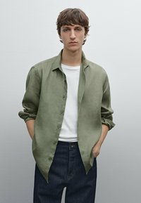 Lichtgroene linnen button-up shirt, lange mouwen opgerold tot aan de ellebogen, gedragen over een wit T-shirt, gecombineerd met een donkerdenim jeans. Neutrale achtergrond.