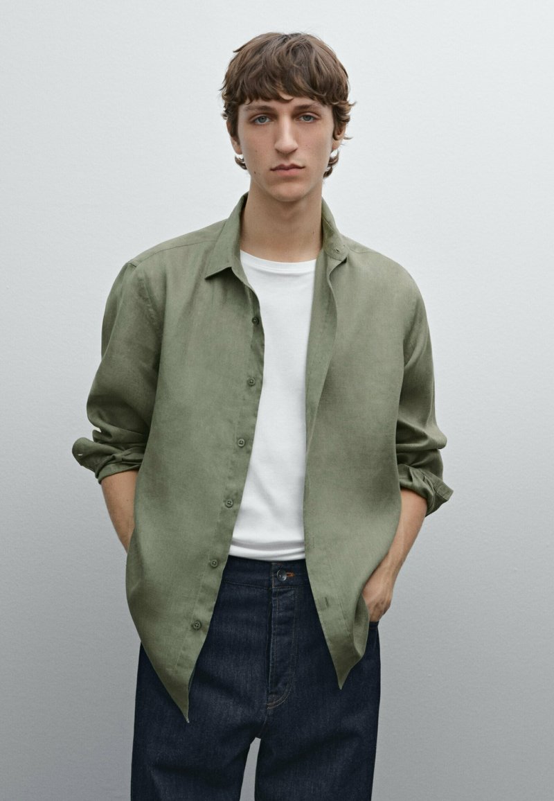 Lichtgroene linnen button-up shirt, lange mouwen opgerold tot aan de ellebogen, gedragen over een wit T-shirt, gecombineerd met een donkerdenim jeans. Neutrale achtergrond.
