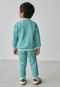 Tyrkysová bavlnená tepláková súprava zahŕňa pulóver s okrúhlym výstrihom a pruhovanými manžetami a zodpovedajúce zúžené nohavice. Biele tenisky s modrými akcentmi.