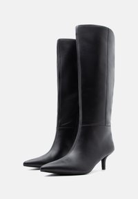 Par crnih kožnih čizama do koljena s uskim vrhovima i tankim niskim potpeticama na jednobojnoj bijeloj pozadini.