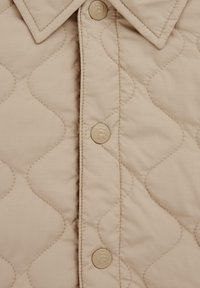 Beige kankainen kangas, jossa on timanttimuotoilu, varustettuna kärjellä kauluksella ja nappikiinnityksellä, jonka napit ovat pyöreitä beigejä.