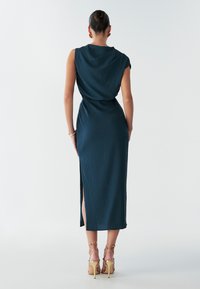 Robe bleu sarcelle arrivant au genou, en tissu texturé, avec des manches courtes, taille cintrée et fente sur le côté. Portée avec des talons légers à lanières.