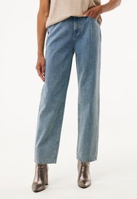 Jeans de mezclilla de corte ancho en un tono azul claro, con un patrón sutil moteado, que presentan una cintura alta y un corte recto, combinados con botas metálicas hasta el tobillo.