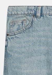 Jeans in denim blu chiaro con tasca anteriore, cuciture visibili e un bottone metallico decorativo. La texture appare leggermente sbiadita.
