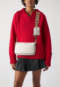 Borsa a tracolla a scacchi beige con dettagli dorati, dotata di una piccola pochette staccabile. Indossata sopra un maglione a maglia rosso e una gonna a quadri neri.