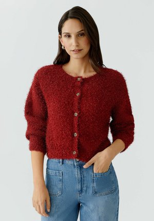 Femme portant un cardigan rouge à boutons, duveteux, à manches longues, associé à un jean taille haute bleu avec des poches plaquées à l'avant.