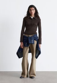 Brown ribbed long-sleeve top, fitted with a collar and button detail; paired with wide beige trousers and a denim jacket tied at the waist.