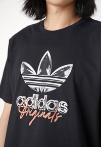 Černé bavlněné tričko s metalickým stříbrným logem Adidas a designem listu, pod kterým je v červeném písmu napsáno "Originals".
