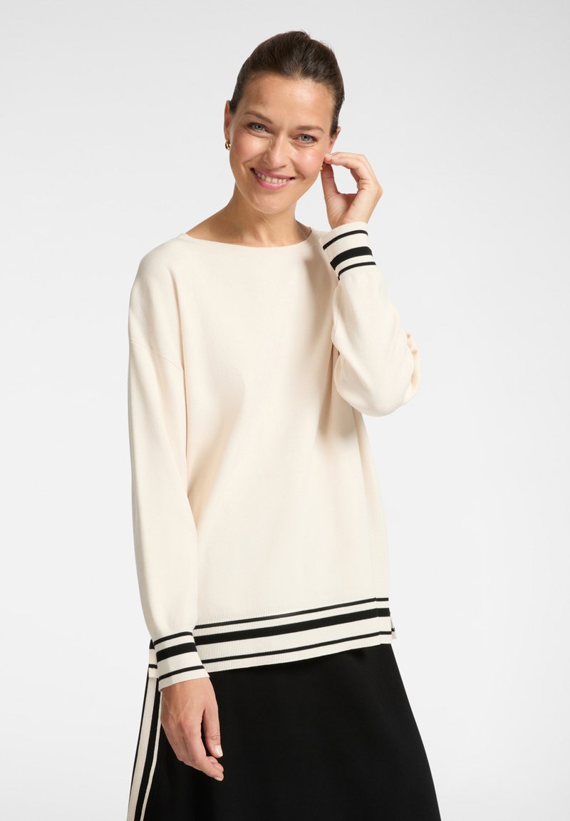 Frau lächelt, trägt einen cremefarbenen Pullover mit schwarzen Streifen an den Bündchen und am Saum, kombiniert mit einem schwarzen Rock, vor einem einfarbigen hellen Hintergrund.