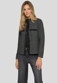 Graue gewebte Jacke mit strukturierter Passform, schwarzen Paillettenakzenten an der Vorderseite und zwei vorderen Taschen. Kombiniert mit dunklen Jeans.