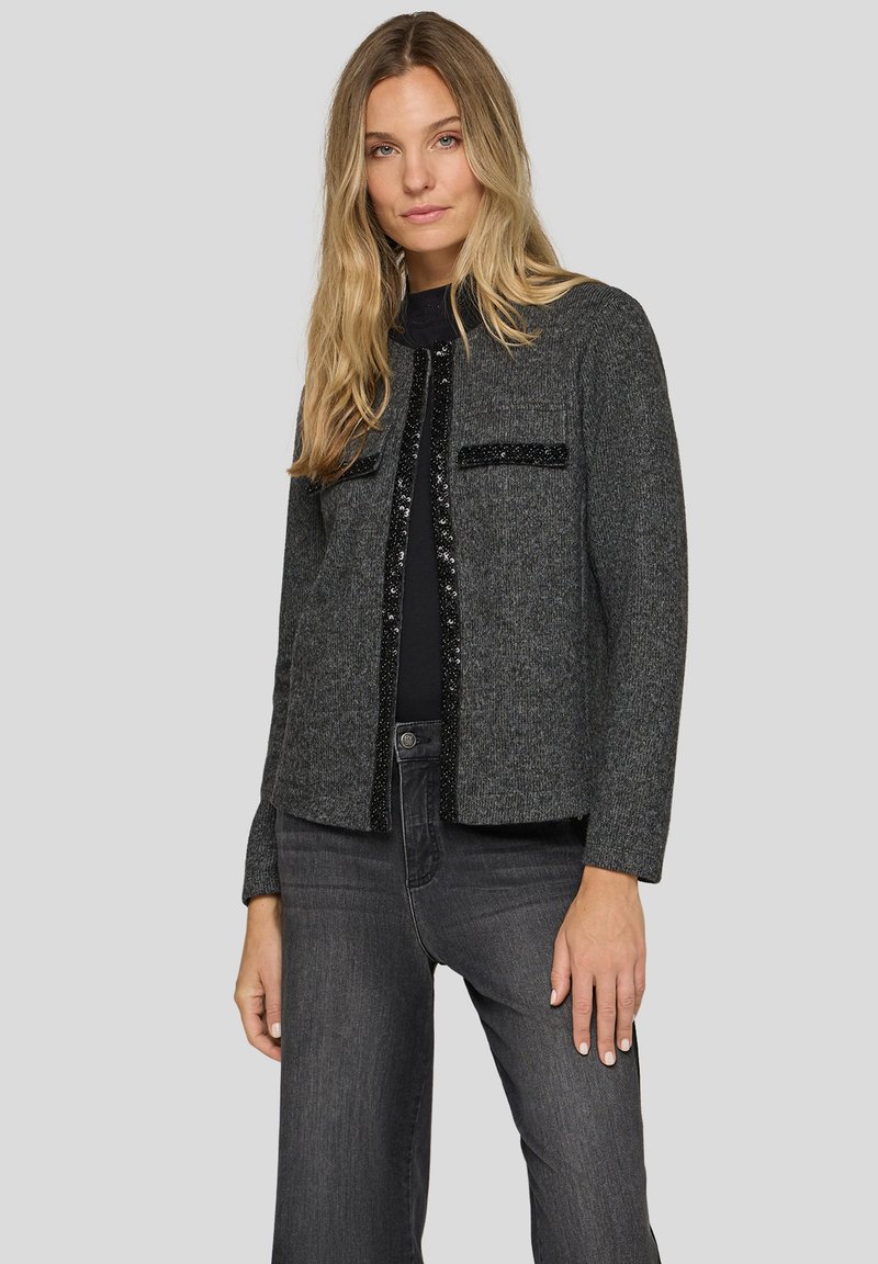 Graue gewebte Jacke mit strukturierter Passform, schwarzen Paillettenakzenten an der Vorderseite und zwei vorderen Taschen. Kombiniert mit dunklen Jeans.