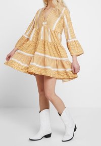 Gelbes Kleid mit Muster, langen Glockenärmeln, V-Ausschnitt, gestuftem Rock und weißen Akzenten; kombiniert mit weißen Cowboystiefeln.