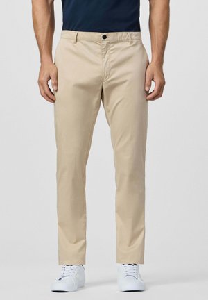 Homme portant un pantalon beige droit, un haut bleu marine et des baskets blanches, debout devant un fond clair uni.