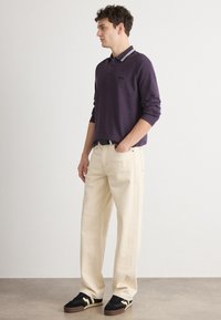 Μωβ μακρυμάνικο πουκάμισο πόλο με λευκές λεπτομέρειες στον γιακά, συνδυασμένο με κρεμ χρώματος παντελόνι σε φαρδιά γραμμή και μαύρα αθλητικά παπούτσια με μπεζ λεπτομέρειες.