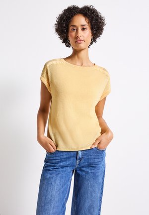 Top jaune en tricot à manches courtes avec une finition texturée, col rond et coutures d'épaule tombantes, associé à un jean bleu à jambes larges.