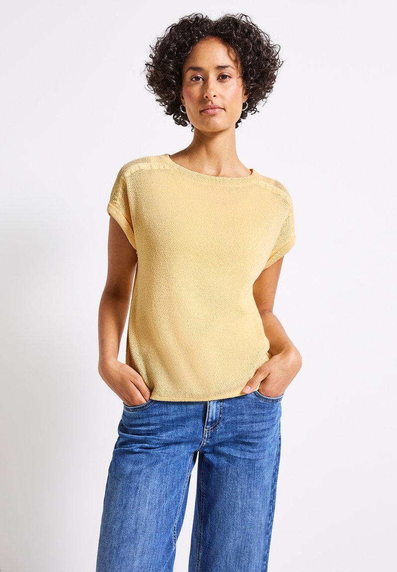 Gul stickad kortärmad topp med en strukturerad yta, rund halsringning och sänkta axelsömmar, parat med blå vida jeans.