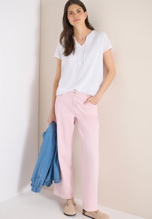 Kvinde iført hvid kortærmet top og lyserøde bukser, der holder en blå denimjakke, stående foran en beige og hvid baggrund.