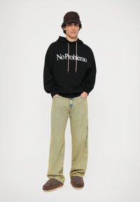 Černá mikina s textem "No Problemo", šedé pruhované kalhoty a hnědé vyšívané pantofle. Hnědá čepice dotváří ležérní outfit.