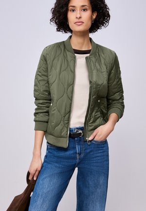 Jeune femme aux cheveux courts et bouclés portant une veste matelassée vert olive, une chemise beige, un jean bleu, une ceinture noire, tenant un sac marron, debout.