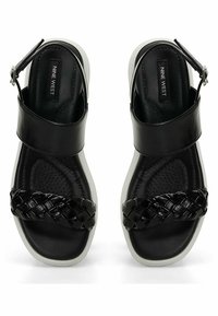 Sandalias negras con una tira trenzada, material similar al cuero, plantilla acolchada y correa ajustable en el tobillo. La suela blanca añade contraste.