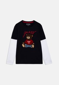 Schwarzes T-Shirt mit weißen langen Ärmel, versehen mit einem Grafik eines braunen Bären in Jeanskleidung und rotem Text "guess" in geschwungener Schrift.