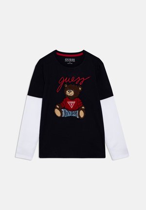 Schwarzes T-Shirt mit weißen langen Ärmel, versehen mit einem Grafik eines braunen Bären in Jeanskleidung und rotem Text "guess" in geschwungener Schrift.