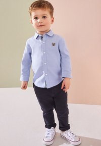 Jeune garçon aux cheveux châtain clair portant une chemise bleu clair boutonnée avec un écusson ours, un pantalon foncé et des baskets blanches, debout à l'intérieur.