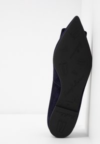 Chaussure plate de ballet pointue bleu marine à semelle noire, debout sur le talon sur fond blanc, montrant des icônes embossées de marque et de chaussons de ballet.