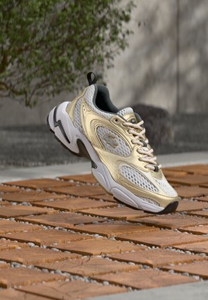 Λευκό και μπεζ παπούτσι με λεπτομέρειες από δίχτυ και δέρμα, αιωρούμενο πάνω από καφέ ξύλινη επιφάνεια με θολό πράσινο φυτό και γκρι τοίχο στο φόντο.